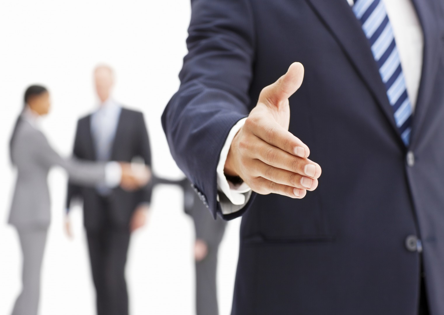 A businessman is seen extending his hand in greeting while other businesspeople stand in the background. Horizontal shot.